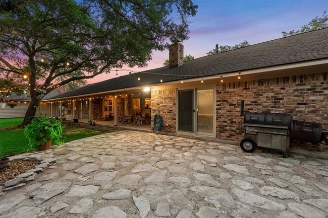 This charming backyard features a spacious stone patio, ideal for outdoor gatherings. It includes a cozy covered porch with warm string lights, a brick exterior, lush greenery offering a perfect blend of comfort and style for entertaining. | Image 28