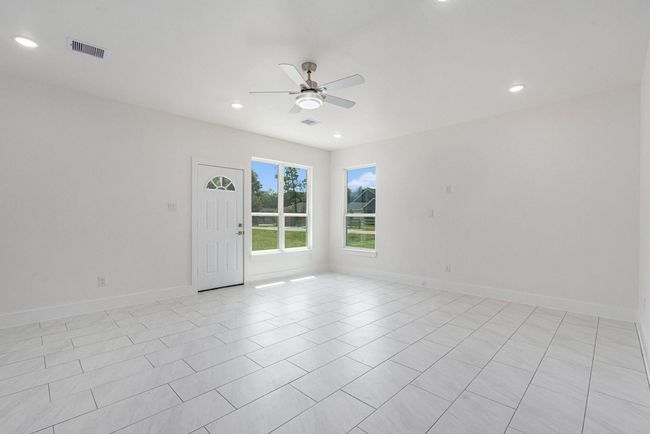 This photo shows a bright, spacious living area with light-colored tile flooring, a ceiling fan, recessed lighting, and large windows providing plenty of natural light. The neutral walls create a clean, modern look. | Image 6