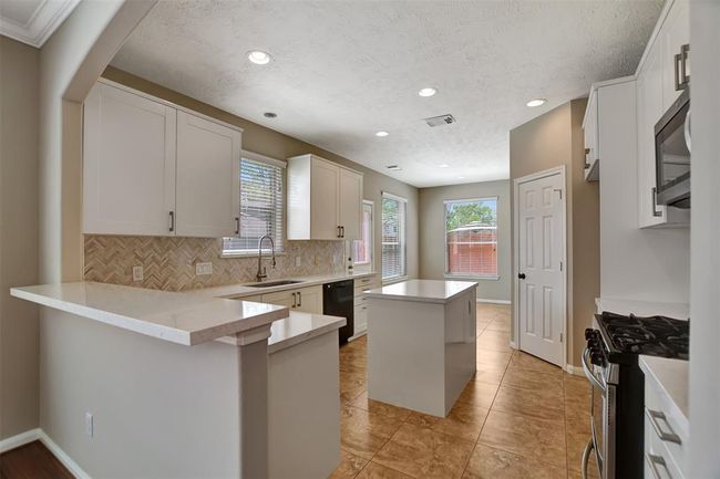 This modern kitchen features sleek white cabinetry, a central island, and stainless steel appliances. The space is brightened by large windows and recessed lighting, with a tiled backsplash adding an elegant touch. Ideal for cooking and entertaining. | Image 20