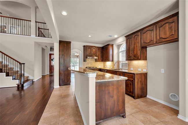 Spacious kitchen with rich wooden cabinets, granite countertops, and a central island. It opens to a hallway with a staircase featuring elegant iron railings and hardwood floors, creating a warm and inviting atmosphere. | Image 12