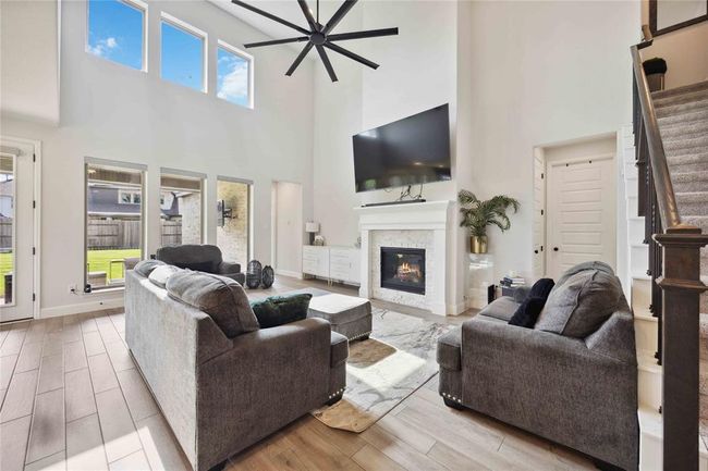 This spacious living room features high ceilings, abundant natural light from large windows, a modern ceiling fan, and a cozy fireplace with a mounted TV above. It offers a stylish, comfortable setting with plush seating and a view into the backyard through tall glass doors. | Image 10