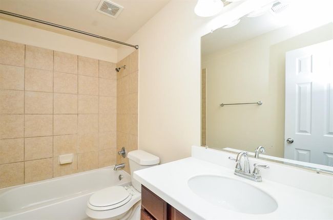 This secondary bathroom features a clean, modern design with beige tiled walls surrounding a bathtub, a white vanity with a sleek faucet, and a large mirror. The space is well-lit and includes a towel rack and standard fixtures. | Image 28