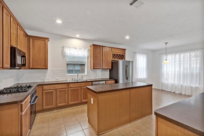 This bright kitchen features wooden cabinetry, a central island, stainless steel appliances, and a gas stove. It includes ample counter space, large windows with sheer curtains, and an open dining area with tile flooring. | Image 7