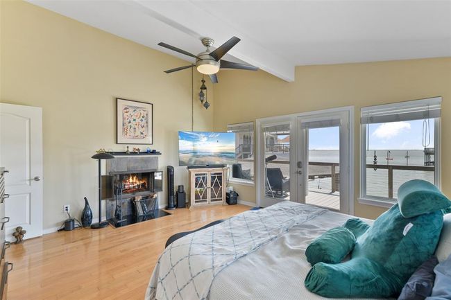 This spacious primary bedroom features the other half of the double sided fireplace, a ceiling fan, and large windows offering stunning waterfront views. The room opens to a deck, providing easy access to outdoor relaxation. | Image 26