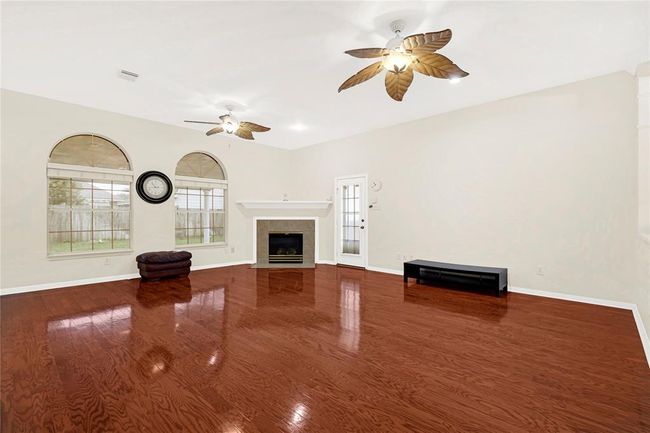 Spacious living room with polished hardwood floors, a central fireplace, and large arched windows offering plenty of natural light. The room features ceiling fans and neutral walls, creating a warm and inviting atmosphere. | Image 6