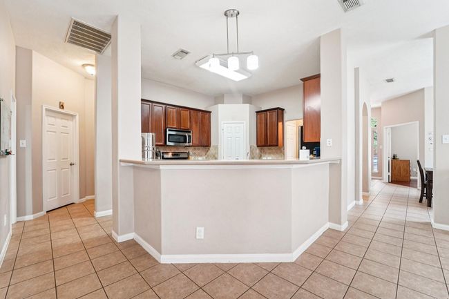 This kitchen features an open layout with a central island, modern lighting, and ample cabinetry. It includes stainless steel appliances and tile flooring, seamlessly connecting to adjacent living areas for easy entertaining. | Image 10