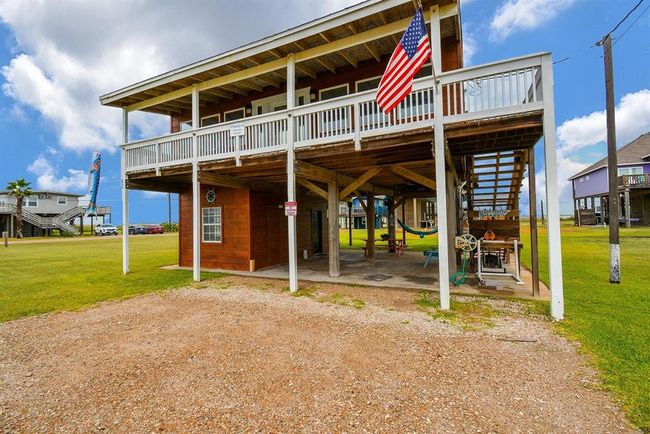 This elevated beach house features a spacious wraparound deck, an American flag, and ample covered parking beneath. It's set on a grassy lot with nearby homes, offering a coastal, relaxed vibe. | Image 32