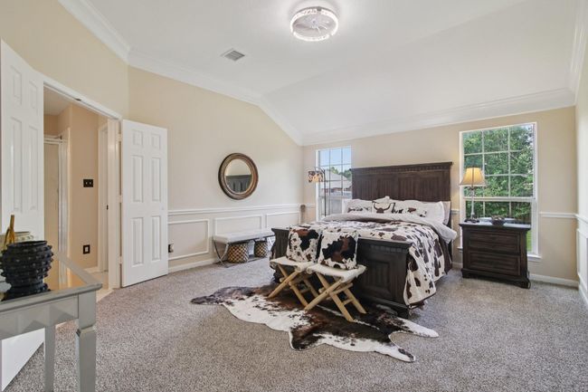 This spacious bedroom features soft carpeting, neutral walls, and abundant natural light from large windows. It includes elegant moldings, and a stylish ceiling fan. | Image 16