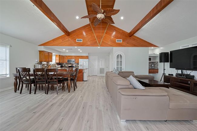 Spacious open-concept living area features a vaulted ceiling with wood accents and a dining area that flows into a modern kitchen with wooden cabinets and stainless steel appliances. New laminate flooring and neutral tones create a warm, inviting atmosphere. | Image 5