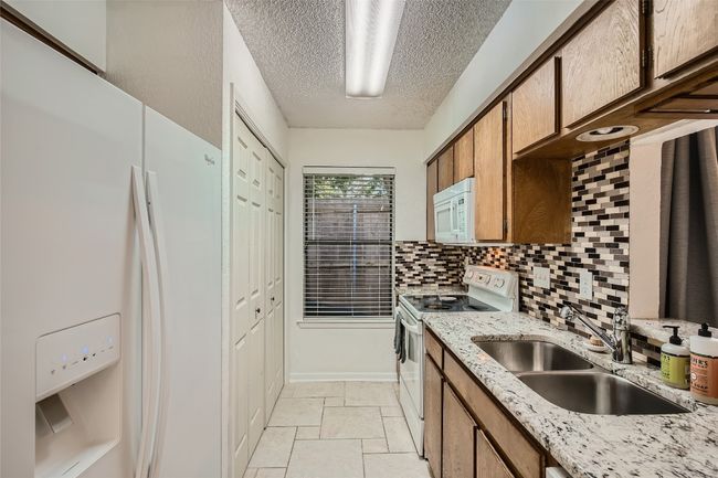 Kitchen with white appliances, a textured ceiling, brown cabinetry, backsplash, and stone tile floors | Image 6