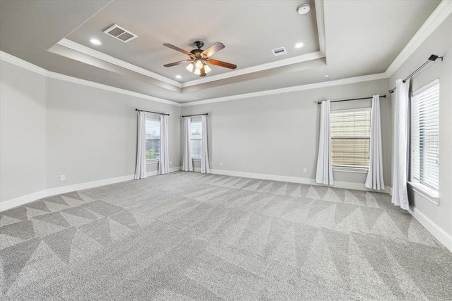 Spacious, light-filled primary bedroom with plush carpeting, neutral walls, and a tray ceiling featuring a central fan. Natural light streams through multiple windows adorned with white curtains, creating an inviting atmosphere. | Image 14