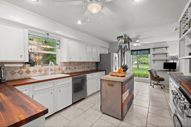 This kitchen features white cabinetry, butcher block countertops, and a central island with a rustic touch. It includes stainless steel appliances, tiled flooring, and a cozy workspace by a large window, creating a bright and inviting atmosphere. | Image 11