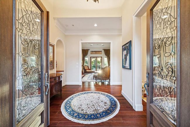 This welcoming entryway features elegant double doors with intricate glass designs, leading to a warm, hardwood-floored hallway. A decorative round rug adds charm, and the space opens into a bright living area with large windows and tasteful furnishings, creating an inviting atmosphere. | Image 7