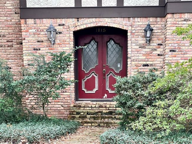 Charming brick exterior with a vintage feel, featuring a unique red double door with decorative glass panels. Flanked by classic lantern-style lights and framed by lush greenery, the entrance exudes a warm, welcoming ambiance. | Image 4