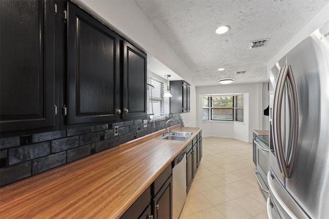 This kitchen features sleek black cabinetry, a modern stainless steel refrigerator, and a double sink. It has a bright, open layout with a cozy breakfast nook by large windows, providing ample natural light. | Image 16