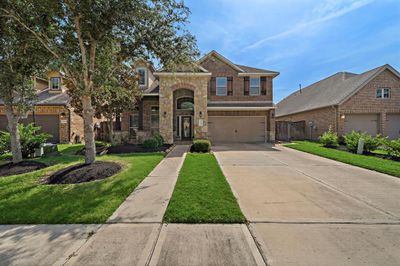 This is a two-story brick home featuring stone accents around the entrance, a two-car garage, and a well-maintained front yard with mature trees. | Image 2