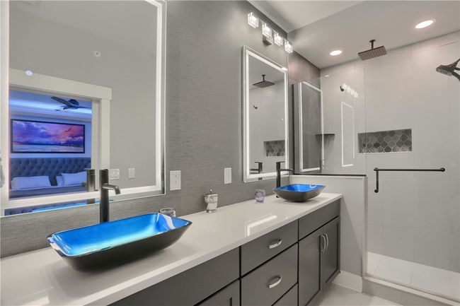 This stunning primary bathroom features dramatic backlit mirrors, bold blue vessel sinks, sleek matte black fixtures, and a spacious walk-in shower with rainfall head and geometric tile detailing—delivering spa-like luxury with a modern edge. | Image 34