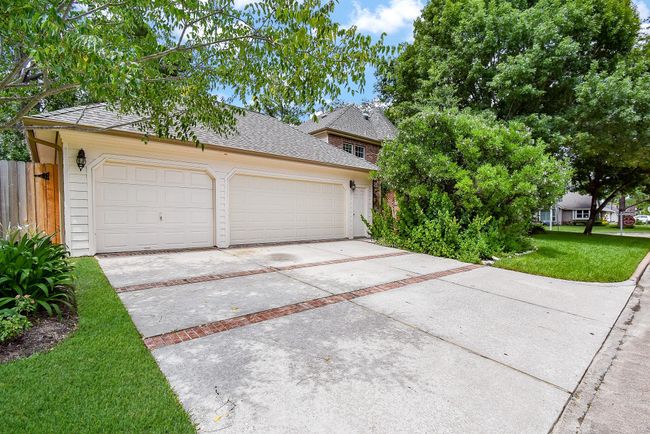 This photo shows a spacious driveway leading to a two-car garage attached to a brick home, surrounded by lush greenery and trees, in a suburban setting. | Image 43