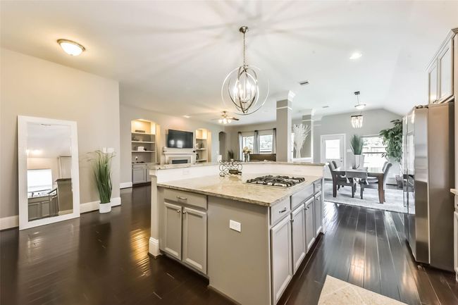 A modern, open-concept kitchen and living area with dark hardwood floors. The kitchen features a central island with a gas cooktop, stainless steel appliances, and stylish pendant lighting. The space flows seamlessly into a dining area with ample natural light, highlighting the home's contemporary design and inviting atmosphere. | Image 13