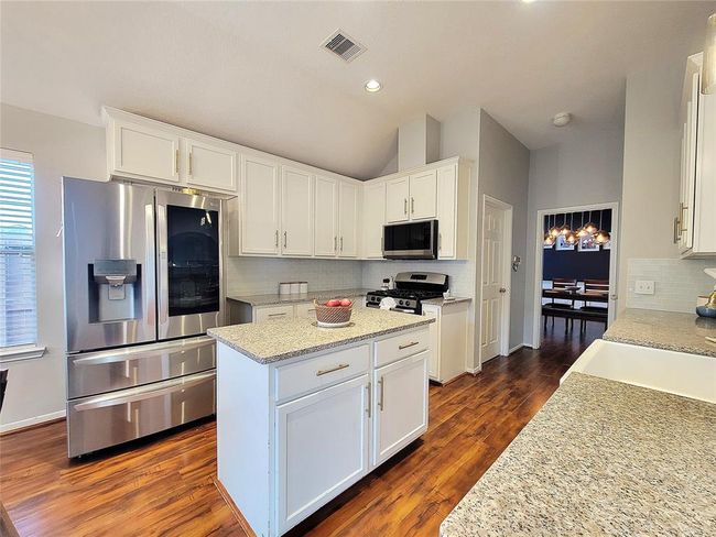 This kitchen features modern stainless steel appliances, white cabinetry, and granite countertops. It has a central island, wood flooring, and an open layout leading to a dining area with stylish lighting. Refrigerator stays! | Image 9