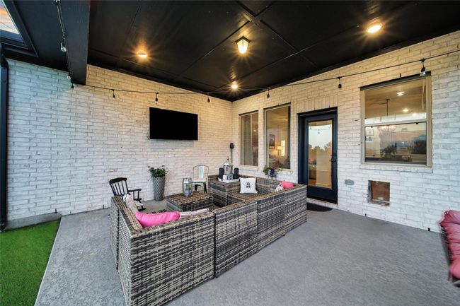 This inviting patio features a cozy wicker seating area with pink cushions, a mounted TV, and overhead lighting. It's complemented by white brick walls, a black ceiling, and a door leading inside. Perfect for relaxing or entertaining. | Image 41