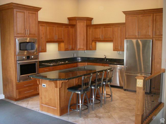 Kitchen with appliances with stainless steel finishes, brown cabinetry, a kitchen island, a breakfast bar, and dark stone countertops | Image 17