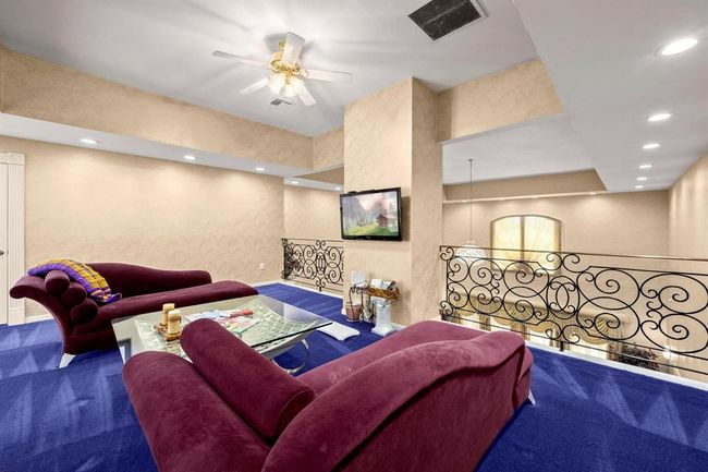 This is a spacious, primary bedroom, balcony area featuring plush maroon sofas, a glass coffee table, blue carpeting, and a decorative wrought iron railing. The room includes recessed lighting, a ceiling fan, and a mounted flat-screen TV. | Image 14