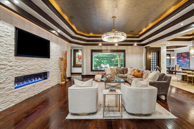 This spacious living room blends comfort and sophistication, featuring a dramatic tiered ceiling with cove lighting and a cork texture ceiling detail. A sleek linear fireplace with crushed marble stacked stone surround adds warmth and ambiance beneath a built-in media display. Rich, hand scraped wood floors flow seamlessly into the open-concept layout, while oversized plate-glass windows frame serene views of the covered outdoor living area and greenspace beyond. | Image 12