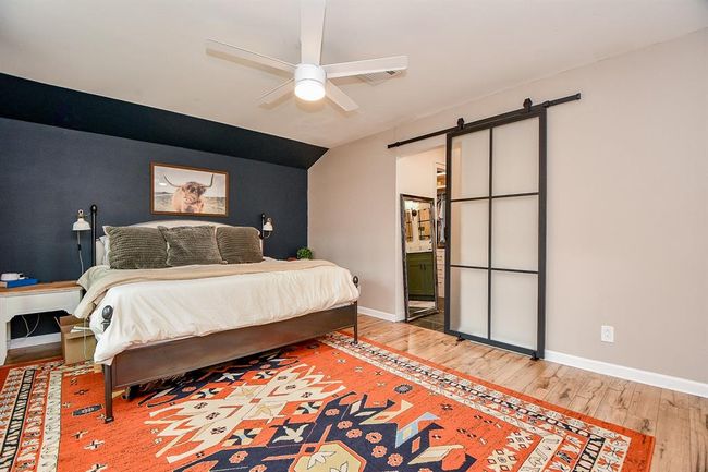 This bedroom features a cozy, modern design with a large bed against a dark accent wall. It has a stylish ceiling fan, wood flooring, and a vibrant patterned rug. A sliding barn door leads to an adjacent space, adding a contemporary touch. | Image 26