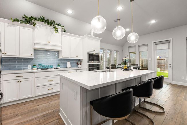 This stunning kitchen displays such a beautiful contrast with the white cabinets, neutral paint, quartz countertops, blue modern backsplash, SS appliances, oversized kitchen island with extra storage, breakfast bar, recessed lighting, high ceilings and modern pendants over the bar. | Image 5