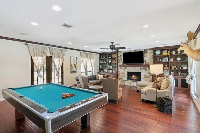This spacious living area features rich hardwood floors, a cozy stone fireplace, and built-in shelves. It includes a pool table for recreation, comfortable seating, and large windows for natural light. Perfect for entertaining and relaxation. | Image 13