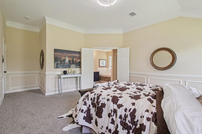 This spacious bedroom features neutral walls with elegant wainscoting, plush carpeting, and a tray ceiling. Double doors open into an adjoining ensuite bath, enhancing the open feel. | Image 17
