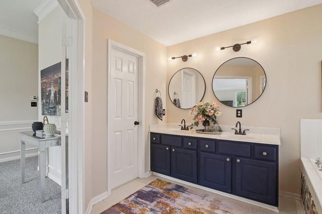 This bathroom features a modern double vanity with navy cabinets, round mirrors, and stylish lighting. The neutral walls and decorative accents create a welcoming atmosphere, and there's a glimpse of the primary bedroom with carpeted flooring. | Image 20