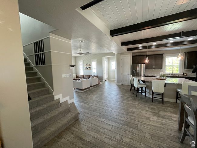 Great room featuring a living room with a ceiling fan, chefs kitchen, wood-style tile flooring, beam/shipped lapped ceiling, and staircase leading upstairs | Image 9