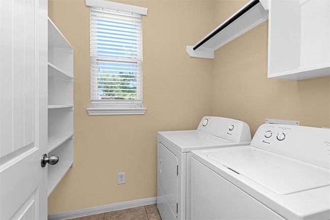 The spacious laundry room is thoughtfully designed to accommodate full-size appliances with ease. An overhead shelf with a hanging rod provides a convenient spot for drying delicates & storing laundry essentials. Opposite the machines, built-in shelving adds more storage space. | Image 11
