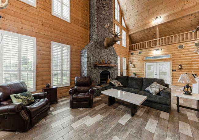 Living area with a towering ceiling, wood look tile floors, a stone fireplace, and log walls | Image 5