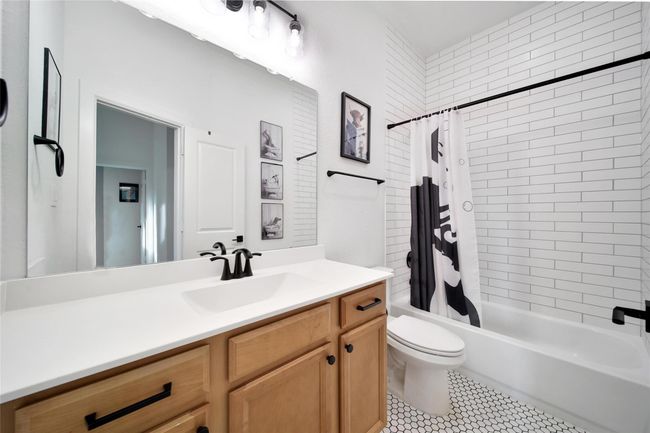 Modern bathroom featuring a sleek vanity with wood cabinetry, a large mirror, and black fixtures. It includes a bathtub with white subway tile surround and a stylish shower curtain. Hexagonal tile flooring adds a classic touch. | Image 35