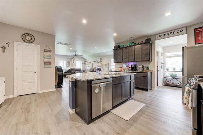 Kitchen featuring appliances with stainless steel finishes, dark brown cabinetry, light wood finished floors, light stone counters, and an island with sink | Image 19