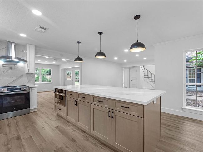 Modern kitchen with an open layout, featuring a spacious island with pendant lighting. It has wood flooring, sleek cabinetry, and stainless steel appliances. Large windows provide natural light, and there's a view of the adjacent living area. | Image 14
