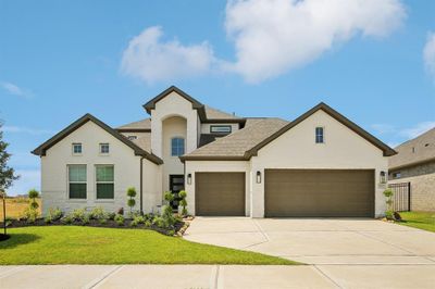 This is a modern two-story home featuring a brick facade, with a prominent central entrance flanked by large windows. It includes an attached three-car garage and a neatly landscaped front yard. The residence has a combination of gable and hip rooflines, and it's situated under a clear blue sky, suggesting a pleasant suburban setting. | Image 1