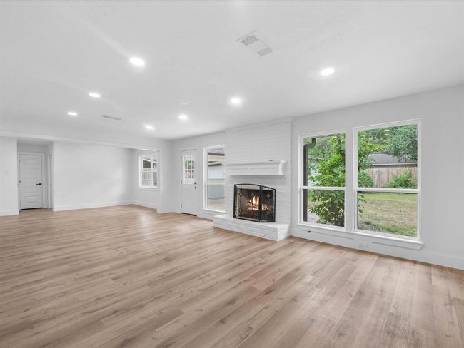 This spacious living room features light wood flooring, a cozy white brick fireplace, and large windows offering plenty of natural light and views of the backyard. The open layout provides ample space for various seating arrangements. | Image 7