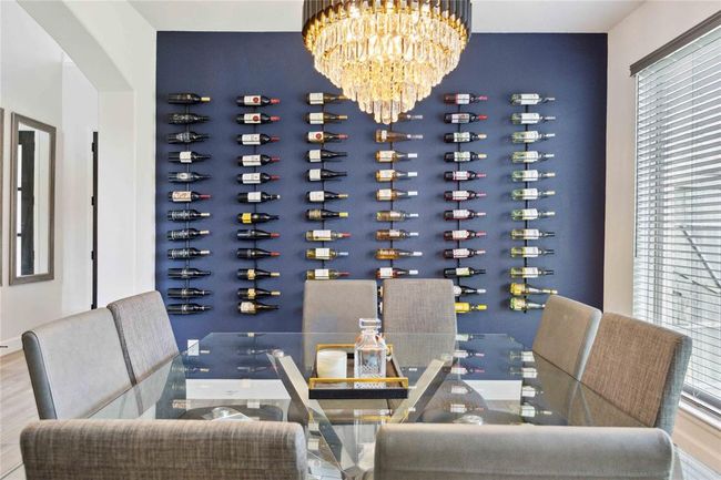 This elegant dining room features a striking wall-mounted wine display, modern gray upholstered chairs, and a glass-top table. A luxurious chandelier adds a touch of sophistication, while large windows provide ample natural light. | Image 7