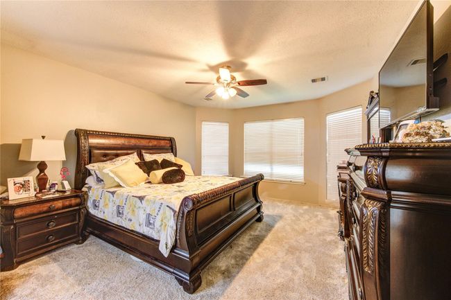 This cozy bedroom features plush carpeting, a ceiling fan, and ample natural light from multiple windows. It includes elegant dark wood furniture with a large bed, dresser, and nightstand, creating a warm and inviting atmosphere. | Image 21
