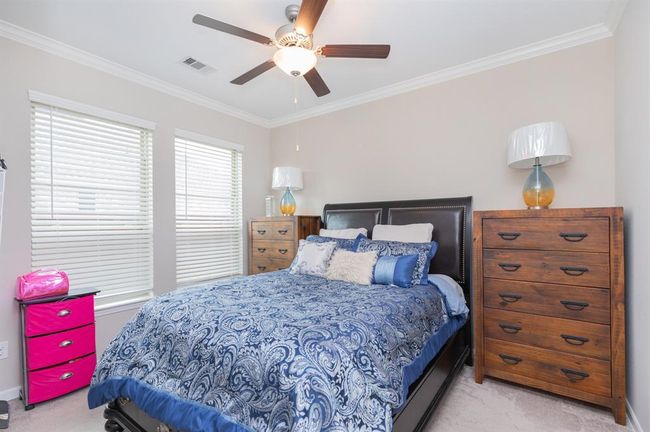 This cozy bedroom in the Next Gen suite offers a peaceful retreat with neutral walls, carpeted flooring, and a ceiling fan for added comfort. A large bed with a blue patterned comforter is complemented by two wooden dressers, providing ample storage space. The room is bathed in natural light from two windows, creating a serene atmosphere perfect for rest and relaxation. | Image 32