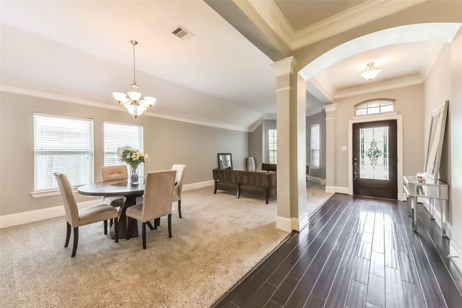 The spacious, open-concept living and dining area features elegant crown molding, soft carpet flooring, and ample natural light from large windows. A stylish chandelier highlights the dining space, creating a warm and inviting atmosphere. Perfect for entertaining and comfortable family living. | Image 5