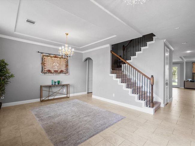 This elegant entryway features a stylish staircase with wrought iron railings, a decorative chandelier, and neutral tile flooring. An arched doorway and a light gray color palette enhance the spacious, inviting atmosphere. | Image 40