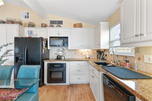 This kitchen features white cabinetry and granite countertops, with a modern black refrigerator and oven. It includes a built-in microwave, dishwasher, and ample natural light from a window above the sink. The space is accented with neutral tile backsplash and hardwood flooring, creating a warm and inviting atmosphere. | Image 17