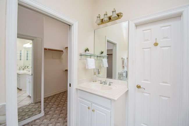 This photo shows a bathroom with a single sink vanity, a large mirror, and a three-light fixture above. There are white cabinets and a door leading to another room. The space is bright and clean, with neutral tones. | Image 30
