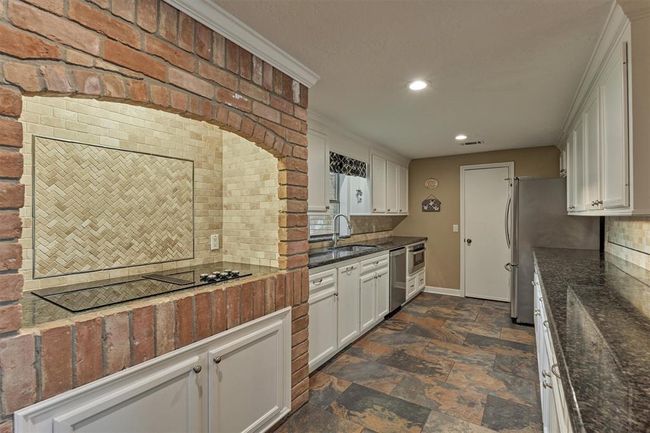 The kitchen features a unique brick archway over the cooktop, complemented by a herringbone tile backsplash. Sleek granite countertops, white cabinetry, and stainless steel appliances complete the space, while slate-look tile flooring adds warmth and character. | Image 17