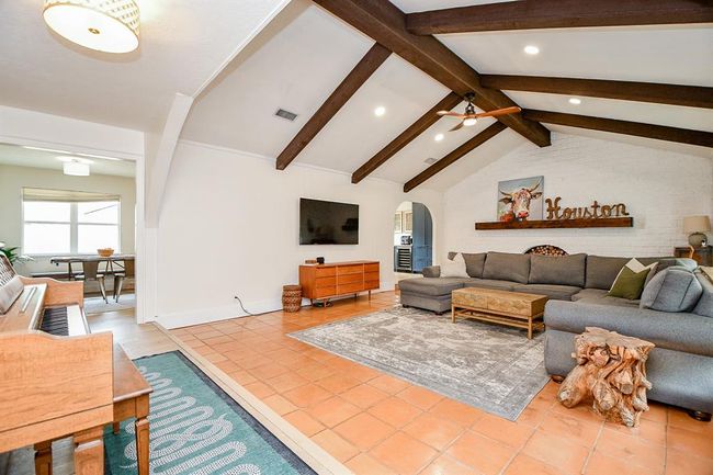 Spacious living room featuring exposed wooden beams, a cozy sectional sofa fits, and a rustic accent table. The area is well-lit with recessed lighting, and a large wall-mounted TV adds modern convenience. A piano and adjoining dining space enhance the open layout. | Image 12